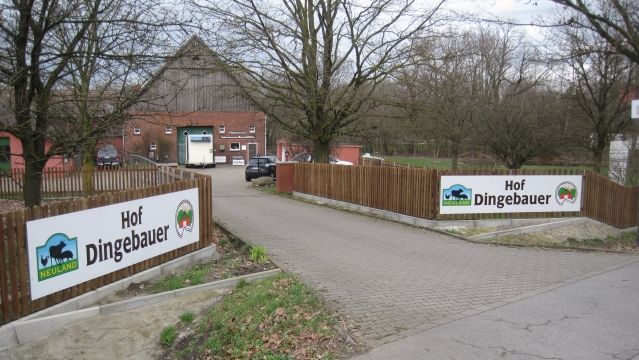 Einfahrt zum Neuland-Hof Dingebauer. Am Zaun hängen zwei Banner mit dem Hofnamen und dem Hoflogo 